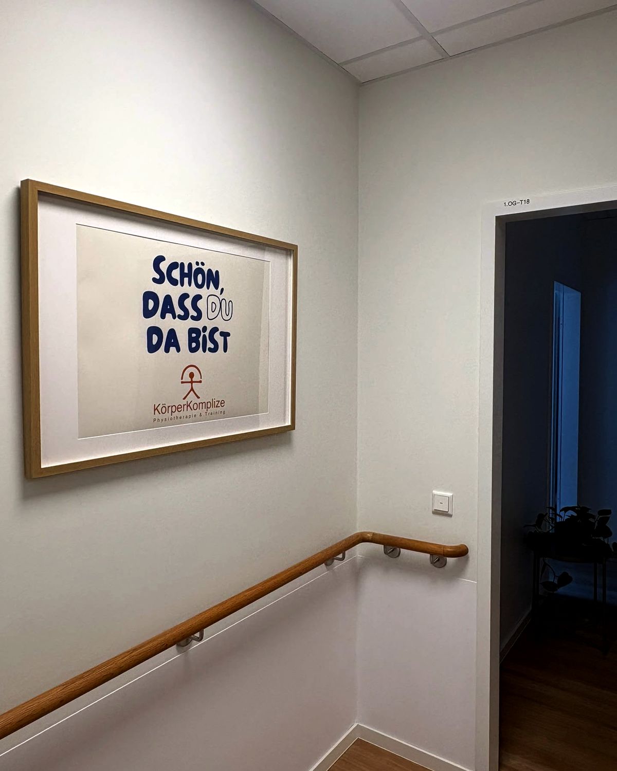 Treppenhaus im ersten Obergeschoss: ein gerahmtes Plakat mit dem Schriftzug „Schön, dass du da bist“ und dem Logo KörperKomplize an der Wand, daneben der Handlauf und die offene Praxistür.