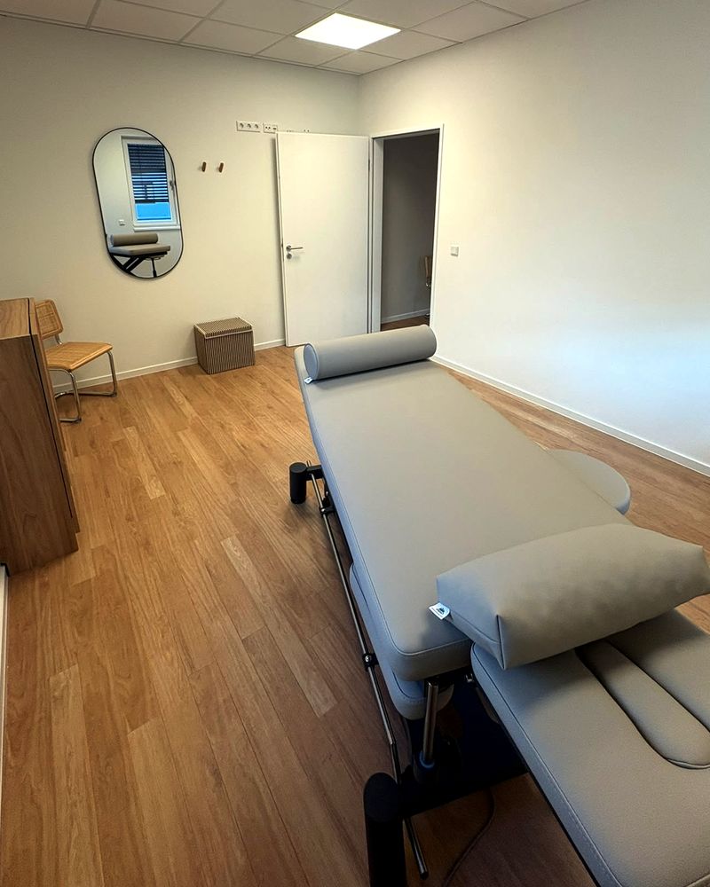 Behandlungsraum mit Therapieliege, Nackenrolle, Wandspiegel, Sitzbank und einem Holzschrank auf hellem Dielenboden.