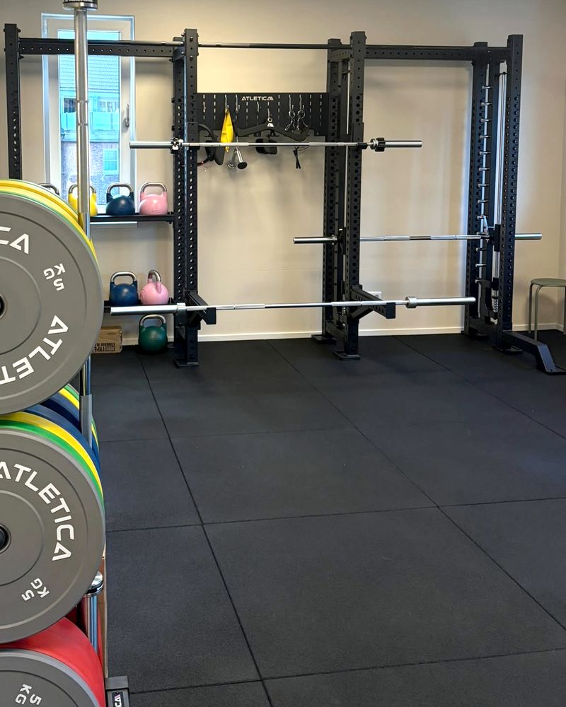 Hantelecke: ein Rack mit zwei Langhanteln, farbige Hantelscheiben auf einem Ständer im Vordergrund und Kettlebells auf einem Wandregal.