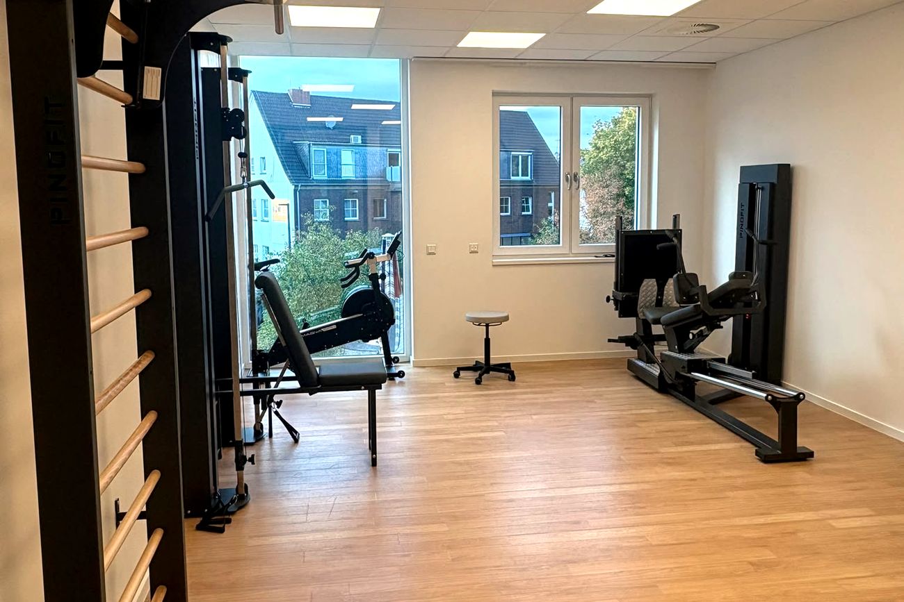 Trainingsfläche der Praxis: Sprossenwand und Seilzug links, Trainingsbank und Ergometer am bodentiefen Fenster, Beinpresse rechts an der Wand.